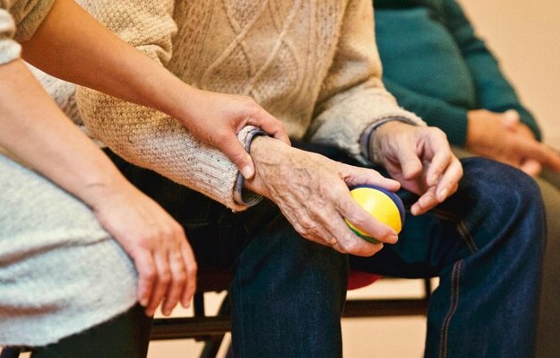 Ein älterer MEnsch hält einen STressball in der Hand, während ihm die Hand eines jüngeren Menschen unterstützend auf dem Arm liegt.