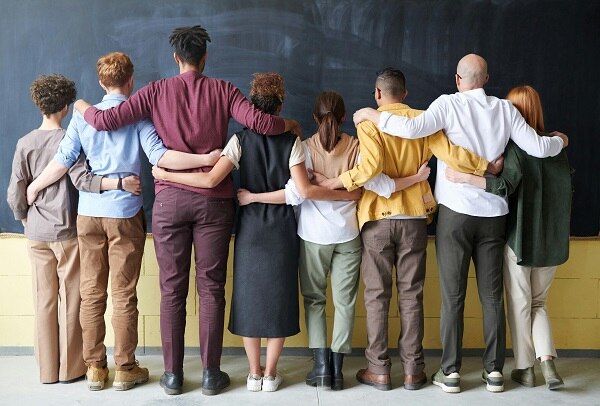 Acht diverse Menschen stehen nebeneinander mit dem Rücken zur Kamera vor einer Tafel und haben die Arme um Schultern bzw. Hüften geschlungen.