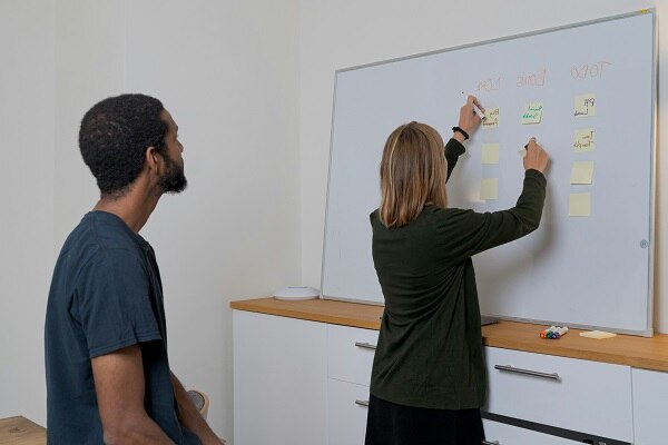 Eine Frau beschriftet Post-Its an einem Whiteboard, ein Mann schaut ihr dabei zu